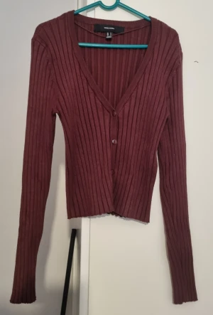 Vinröd ribbad kofta från Vero Moda - Vinröd ribbad kofta från Vero Moda med v-ringning och knappar framtill. Långärmad och figurnära passform, perfekt att styla med jeans eller kjol. Mjuk och stretchig känsla, passar både till lager-på-lager och som den är.