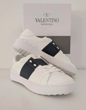 Valentino Garavani vita sneakers - Snygga vita sneakers från Valentino Garavani med bred svart rem över sidan och coola nitar på hälen. Skorna har rund tå, platt sula och snörning framtill. Tillverkade i skinn med stilren design som sticker ut. Perfekta för dig som gillar exklusiva märken och streetstyle.