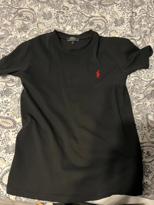 Svart t-shirt från Polo Ralph Lauren - Svart t-shirt från Polo Ralph Lauren med klassisk röd logobroderi på bröstet. Modellen har rund hals och korta ärmar, tillverkad i mjuk bomull som känns skön mot huden. Enkel och stilren design som funkar till allt. Snygg passform och diskret branding.   Pris kan diskuteras..