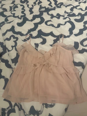 Ljusrosa volangblus med tunna axelband - Supersöt ljusrosa blus med tunna axelband och volangdetaljer framtill och längs sidorna. Blusen är gjord i ett lätt och luftigt material som ger en flowy känsla. Perfekt för varma dagar och har en feminin look med omlott och v-ringad design.