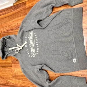 Grå hoodie från Race Marine S - Grå hoodie från Race Marine med vit text på bröstet och stor känguruficka. Tröjan har huva med breda vita snören och är tillverkad i mjuk bomullsmix. Perfekt för chill dagar eller när du vill ha en sportig look.