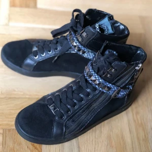 Svarta high-top sneakers Michael Kors - Svarta high-top sneakers från Michael Kors med snörning, dragkedja på sidan och coola blåmönstrade detaljer runt ankeln. Skorna har platt sula, rund tå och är tillverkade i en mix av skinn och mocka. Dekorativa nitar och mönstrad rem ger extra edge.