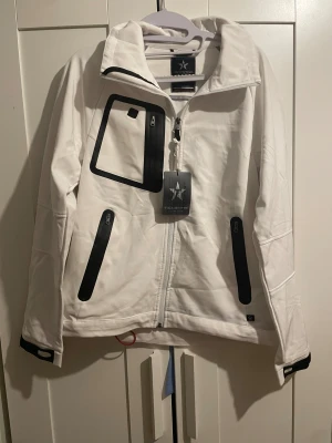 Vit softshelljacka från Texstar - Snygg vit softshelljacka från Texstar med svarta detaljer och dragkedjor. Jackan har hög krage, flera praktiska fickor med dragkedja och justerbara muddar med kardborre. Perfekt för dig som gillar stilren och funktionell design.