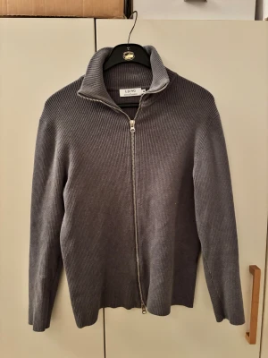 Grå ribbad kofta från Ljung Marcus Larsson - Säljer en grå ribbad kofta från Ljung Marcus Larsson i storlek M. Tröjan har hel dragkedja framtill, hög krage och är långärmad. Materialet är stickat och ger en skön och stilren vibe. Perfekt för dig som gillar minimalistisk design och vill ha något som funkar året runt.