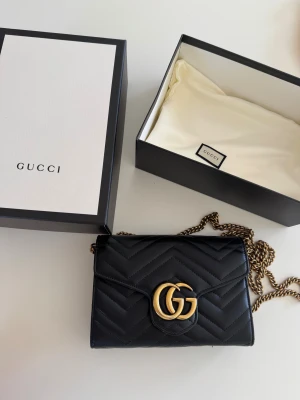 Svart Gucci axelväska i skinn - Snygg svart axelväska från Gucci i mjukt skinn med guldfärgad kedja och GG-logo framtill. Väskan har flera fack och kortplatser samt en dragkedja inuti. 