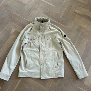 Beige vindjacka från J.Lindeberg M - Stilren beige vindjacka från J.Lindeberg med hög krage, tryckknappar och dragkedja. Jackan har två bröstfickor med lock och knappar samt två sidofickor. Perfekt för dig som gillar minimalistisk och clean look.