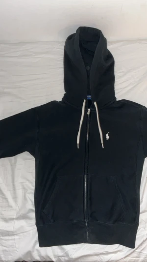 Svart hoodie från Polo Ralph Lauren - Svart hoodie från Polo Ralph Lauren i storlek XS. Tröjan har huva med vita snören, dragkedja framtill och klassisk Polo-logga broderad på bröstet. Mjuk bomullskvalitet och två fickor framtill gör den både chill och snygg.