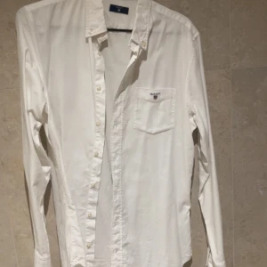 Vit skjorta från Gant med ficka - Klassisk vit skjorta från Gant med lång ärm och bröstficka med broderad logga. Skjortan har knappar hela vägen och en stilren, tidlös design. Perfekt för dig som gillar enkel och clean stil.