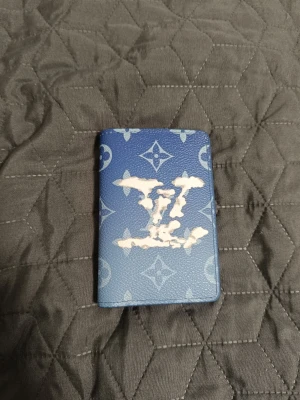 LV Pocket Organizer  - Snygg blå plånbok från Louis Vuitton med klassiskt monogrammönster och stor LV-logga i vitt på framsidan. Tillverkad i skinn med flera kortfack och stilren design. Perfekt accessoar för dig som vill sticka ut med något exklusivt och trendigt.