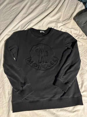 Svart sweatshirt från Moncler - Svart sweatshirt från Moncler med stort, broderat logotypmärke på bröstet. Tröjan har rund hals, ribbade muddar vid ärmslut och nederkant samt långärmad passform. Perfekt för dig som gillar stilrena och exklusiva plagg.