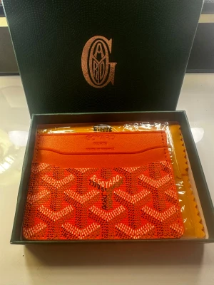 Orange plånbok med mönster - Snygg orange plånbok med ikoniskt grafiskt mönster i vitt och svart. Plånboken har flera kortfack och är tillverkad i exklusivt skinn. Perfekt för dig som vill ha en färgstark accessoar med lyxig vibe och fransk design.