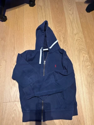 Marinblå hoodie från Polo Ralph Lauren - Snygg marinblå hoodie från Polo Ralph Lauren med dragkedja och vit snörning i huvan. Tröjan har långa ärmar, fickor fram och den klassiska röda Polo-loggan broderad på bröstet. Perfekt för chill dagar och enkel att matcha med jeans.