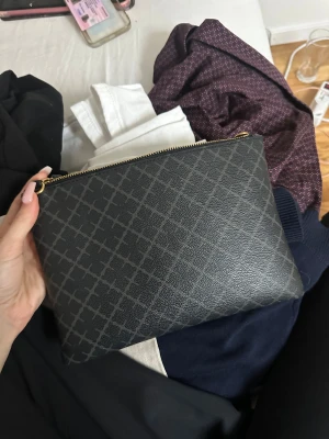 Svart kuvertväska med mönster - Snygg svart kuvertväska med diskret rutigt mönster och guldfärgad dragkedja. Väskan är tillverkad i skinnimitation och har en beige insida. Perfekt storlek för mobilen, plånboken och småsaker. Enkel och stilren design som passar till många outfits. Från By malene birger, band tillkommer inte , Fint skick på utsidan lite smutsig på insidan  band ingår ej