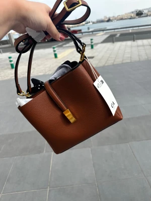 Brun handväska från C&A i skinnimitation - Snygg brun handväska från C&A med stilren design och guldfärgade detaljer. Väskan har justerbar axelrem och ett elegant spänne framtill. Perfekt storlek för det viktigaste och tillverkad i skinnimitation med struktur.