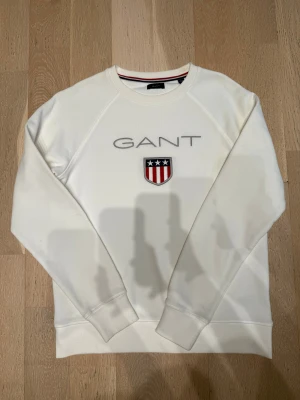 Vit sweatshirt från GANT med broderad logga - Vit sweatshirt från GANT med broderad logga och emblem på bröstet. Tröjan har rund hals, långa ärmar och ribbade muddar. Snygg detalj med röd och marinblå rand på insidan av kragen. Perfekt för en clean och sportig look.