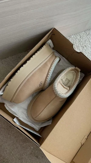 Beige UGG slip-on boots med platåsula - Säljer ett par beige slip-on boots från UGG med platåsula och rund tå. Skorna är i mocka med vit stickad detalj runt öppningen och har mjukt foder inuti för extra komfort. Perfekta för kalla dagar och riktigt trendiga just nu.