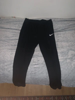 Svarta träningsbyxor från Nike - Svarta mjukisbyxor från Nike med klassisk vit swoosh-logga på låret. Byxorna har en slim passform och är tillverkade i mjukt och stretchigt syntetmaterial, perfekta för träning eller chill. Resår i midjan och smalare benavslut för en modern look. Dessutom är de helt nya! Det står innanför byxorna att storleken är XL men känns snarare som M/L.