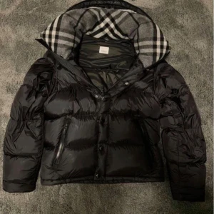 Svart burberry pufferjacka - Snygg svart burberry pufferjacka med glansig finish och rutig huva i grått, vitt och svart. Jackan har dragkedja och knappar framtill samt två sidofickor med dragkedja. Perfekt för kalla dagar och har en cool, urban vibe.