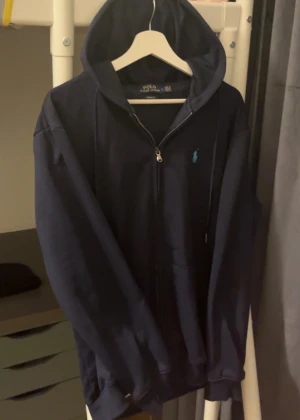 Mörktblåt Polo Ralph Lauren zip hoodie - Säljer en mörkblå hoodie från Polo Ralph Lauren i storlek L. Tröjan har huva med snörning, dragkedja framtill och det klassiska Polo-logot broderat i turkos på bröstet. Materialet är mjuk bomull och passformen är normal. Perfekt för chill dagar.