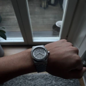 Audemars Piguet Royal Oak silverklocka - Säljer en Audemars Piguet Royal Oak med ikonisk åttakantig boett och länkarmband i borstat stål. Urtavlan är silverfärgad med rutmönster och datumvisning. Baksidan har safirglas som visar det automatiska urverket. Klassisk och stilren design med AP-logga på spännet.