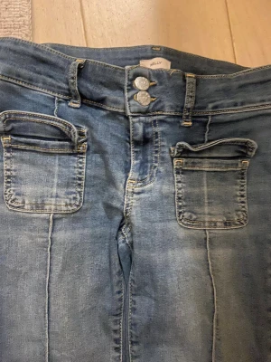 Low waist bootcut jeans från Nelly - Snygga blå jeans från Nelly med bootcut passform och låg midja. Jeansen har dubbla knappar i midjan, markerade sömmar längs benen och stora fickor framtill. Perfekta för en avslappnad och trendig look. Fickorna kan jag få ned med strykjärn och de är långa i långden💕