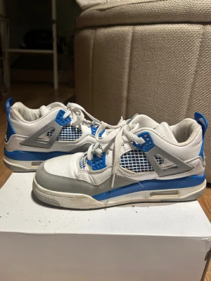 Nike Air Jordan 4 Retro Vit/Blå 38 - Nike Air Jordan 4 Retro sneakers i vitt och blått med grå detaljer. Skorna har meshpaneler, snörning och den klassiska Jumpman-loggan på plösen och hälen. Tillverkade i skinn och syntetmaterial, med platt sula och rund tå. Perfekt för dig som gillar streetwear och ikoniska sneakers.
