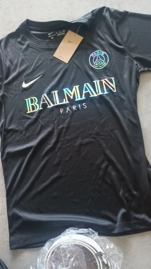 (Äkta) Svart PSG x Balmain fotbollströja - Snygg svart fotbollströja från Nike med Paris Saint-Germain x Balmain Paris tryck i regnbågsskimrande färger. Klassisk rund hals och korta ärmar, tillverkad i lätt och ventilerande polyester. Perfekt för dig som gillar exklusiva samarbeten och streetwear.