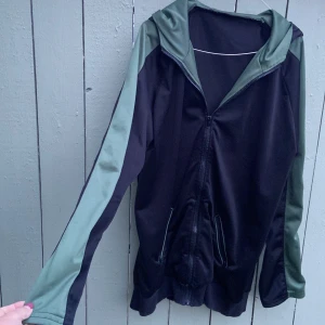 Svart och grön gymnastik jacka med huva - Svart vindjacka gymnastik, träning med gröna detaljer på huva och längs ärmarna. Passar för både dam och herr.Jackan har dragkedja framtill, två fickor med dragkedja och ribbade muddar. Perfekt för dig som gillar sportig stil och vill ha något lätt och smidigt.passar storlek 38-42.mycket bra skick använd 1 gång.