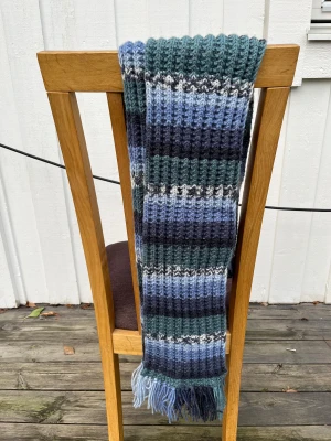 Randig stickad halsduk i blå toner - Mysig stickad halsduk med breda ränder i olika blå nyanser, mörkgrönt, grått och vitt. Ribbstickad struktur och fransar i ändarna. Längd 230cm, bredd 18cm