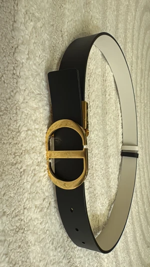 Svart-vitt Dior bälte med gulddetaljer - Snyggt bälte från Dior i svart skinn med vit insida och ikoniskt CD-spänne i guldmetall. Bältet har en stilren och klassisk design som lyfter vilken outfit som helst. Perfekt accessoar för dig som gillar lyxiga detaljer och tidlös stil. Ingen kvitto kvar!