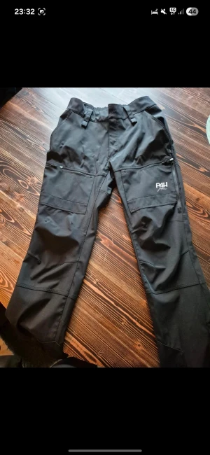 Svarta fritidsbyxor från P4H strl 40 - Svarta byxor från P4H, designade i Sverige. Byxorna har elastisk midja, flera praktiska fickor med dragkedja och är tillverkade i slitstarkt syntetmaterial. Perfekta för utomhusaktiviteter och har en clean, sportig look.