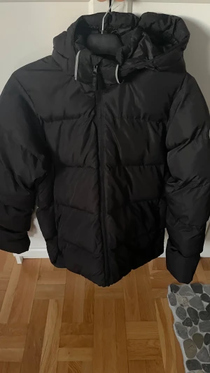 Svart pufferjacka från McKinley 170 - Svart dun jacka från McKinley med huva och dragkedja framtill. Jackan är vadderad och har en enkel, stilren look med reflekterande detalj bak. Perfekt för kalla dagar och riktigt skön att bära. Väldigt fint skick nästan som ny 