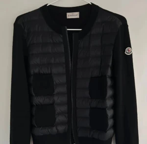 Svart Moncler cardigan  - Säljer en svart cardigan jacka från Moncler med quiltad front och stickade ärmar. Jackan har två fickor framtill, rund halsringning och dragkedja. Klassisk Moncler-logga på ärmen och en clean, stilren look som funkar till allt.