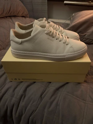 Vita sneakers från Axel Arigato - Snygga vita sneakers från Axel Arigato med stilren design och rund tå. Skorna är tillverkade i slätt skinn och har platt sula samt klassisk snörning. Perfekta för dig som gillar minimalistisk och clean stil. Kommer med originalkartong.