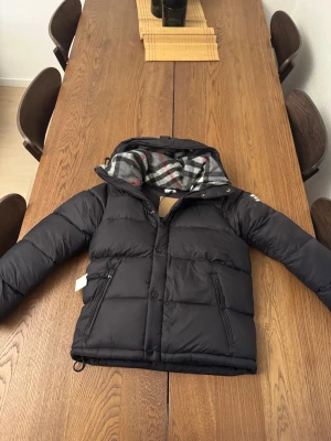 Burberry jacka väst - Svart pufferjacka med huva och rutigt foder i grått, vitt och rött. Jackan har dragkedja och knappar framtill samt två sidofickor med dragkedja. Perfekt för kalla dagar och har en modern, boxig passform.