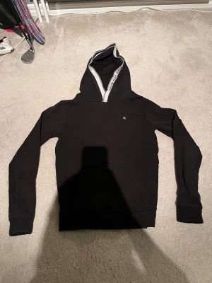 Svart hoodie från Calvin Klein Jeans 14-16 år  - Svart hoodie från Calvin Klein Jeans med vit logga 'ck' på bröstet och vit kant med svart text på huvan. Tröjan har en stor magficka och långa ärmar. Mjuk och skön bomullskvalitet, perfekt för chill dagar. Snygg och enkel design som funkar till allt. Nästan oanvänd 