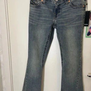 True Religion Joey Low Rise Flare Jeans - Säljer ett par True Religion Joey jeans i modellen Low Rise Flare Flap. De är i en mellanblå tvätt med slitningar och har snygga ficklock bak med dekorativa sömmar och knappar. Jeansen har låg midja och utsvängda ben, tillverkade i klassisk denim. Stl W29. 