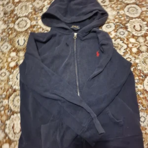 Marinblå hoodie från Polo Ralph Lauren - Säljer en marinblå hoodie från Polo Ralph Lauren med dragkedja och huva. Tröjan har det klassiska röda broderade logotypen på bröstet och är tillverkad i mjuk bomull. Perfekt för chill dagar och snygg till jeans.