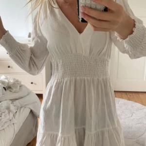 Vit kortklänning med v-ringning Zara - Supersöt vit kortklänning med v-ringad hals och smockad midja. Klänningen har långa ärmar med smock vid ärmslut och volang nedtill. Tillverkad i ett lätt och luftigt bomullstyg, perfekt för varma dagar. Enkel och fräsch stil som är lätt att matcha.