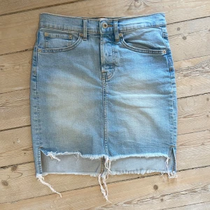 Blå jeanskjol med rå kant - Säljer en ljusblå jeanskjol med klassiska fickor och knappstängning fram. Kjolen har en passform till mitten på låren och rå, fransig kant nedtill som ger en cool och avslappnad vibe. Perfekt för dig som gillar denim och streetstyle.