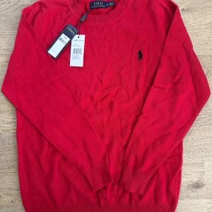 Röd stickad tröja Polo Ralph Lauren - Snygg röd stickad tröja från Polo Ralph Lauren med svart broderad logga på bröstet och RLX-broderi på ärmen. Tröjan har rund hals, ribbade muddar och är långärmad. Perfekt för dig som gillar klassisk stil med en sportig touch.