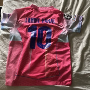 Barça Lamine Yamal #10 rosa matchtröja - Barcelona fotbollströja med Lamine Yamal och nummer 10. Rosa och ljusblå färg med vita detaljer, Nike Dri-FIT material och klubbmärke framtill. Kortärmad modell med tryckta sponsorer och Champions League-märke på ärmen.
