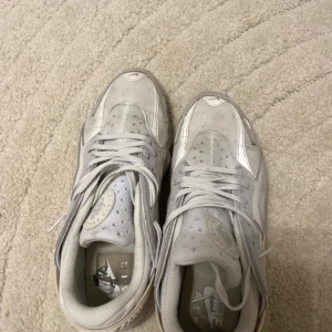 Nike Air Huarache vita sneakers - Nike Air Huarache sneakers i vitt och ljusgrått med klassisk chunky sula. Skorna har mesh, syntet och mockadetaljer samt snörning framtill. Ikonisk logga på plösen och rund tå. Perfekta för dig som gillar retrostil och streetwear.