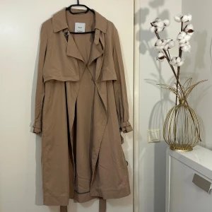 Beige trenchcoat från Mango Suit XS - Snygg beige trenchcoat från Mango Suit i storlek XS. Jackan har klassisk krage, långa ärmar och bälte i midjan för en cool siluett. Tillverkad i mjuk bomull och har detaljer som D-ring och knytband. Perfekt för dig som gillar stilrena och tidlösa plagg.