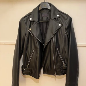 Svart skinnjacka (syntet) från M&S Collection - Snygg svart syntet-skinnjacka från M&S Collection med klassisk bikerstil, silvriga dragkedjor och detaljer vid axlar och midja. Jackan har bred krage, två fickor med dragkedja och spännen i sidorna för extra edge. Perfekt för dig som vill ha en cool och tidlös look.👌👌🤩