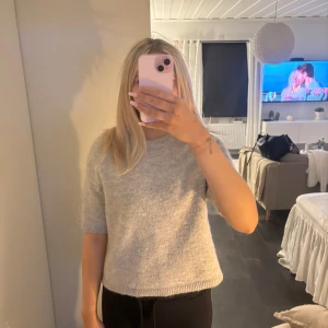 Grå stickad tröja från Gina Tricot - Mysig grå stickad tröja från Gina Tricot med rund halsringning och kortare ärmar. Tröjan har en enkel och clean look, perfekt att styla med jeans eller kjol. Mjuk och skön känsla tack vare det stickade materialet.