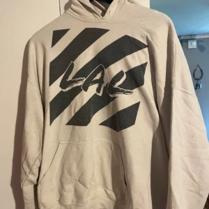 Vit hoodie med svart print LAL - Säljer en beige hoodie med svart grafiskt print och texten LAL på bröstet. Tröjan har huva, känguruficka och är tillverkad i mjuk bomull. Perfekt för dig som gillar streetwear och vill ha en chill look.