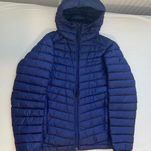 Blå Peak Performance dunjacka med huva - Säljer en blå dunjacka från Peak Performance, modell Frost DH. Jackan har huva, dragkedja framtill och är tillverkad i lätt och slitstark Pertex Quantum. Perfekt för kalla dagar och har en snygg, sportig look.