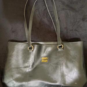 Svart axelväska från Miu Miu - Stilren svart axelväska från Min Min med två långa remmar och guldfärgade ringdetaljer. Väskan är tillverkad i skinnimitation och har ett rymligt brunt innerfoder samt dragkedja upptill. Perfekt för dig som vill ha plats för allt viktigt.