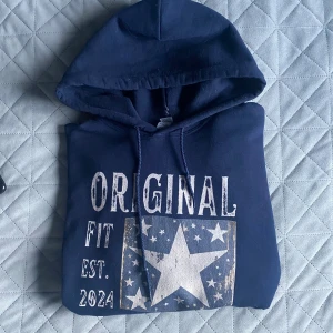Marinblå hoodie från SCNDI - Säljer en marinblå hoodie från SCNDI  i storlek M. Tröjan har ett stort vitt tryck med texten 'ORIGINAL FIT EST. 2024' och en stjärna med mindre stjärnor runtom. Klassisk känguruficka och snörning i huvan. Tillverkad i en mjuk bomulls- och polyestermix.